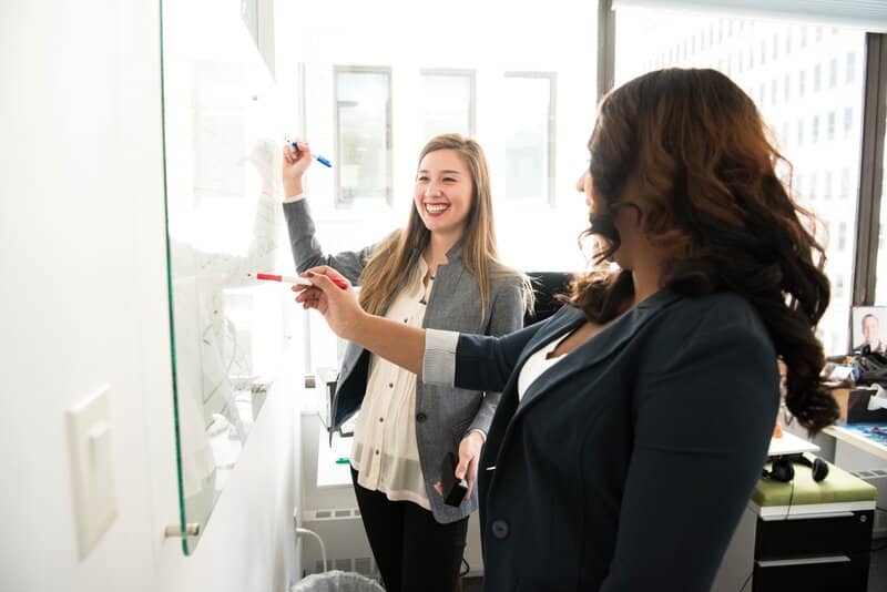 Two women working together writing down a daily office cleaning checklist on a whiteboard.
